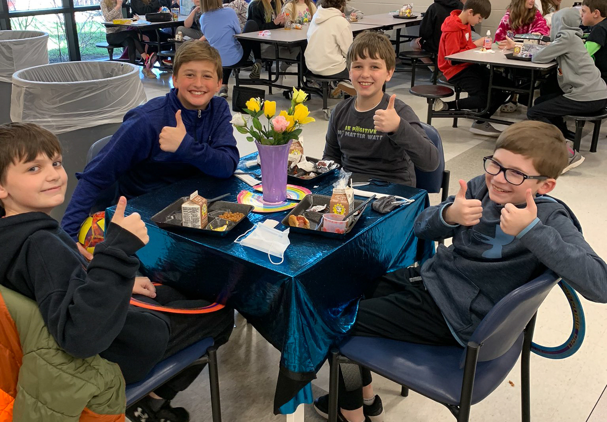 students sitting at a table eat lunch and give the thumbs up